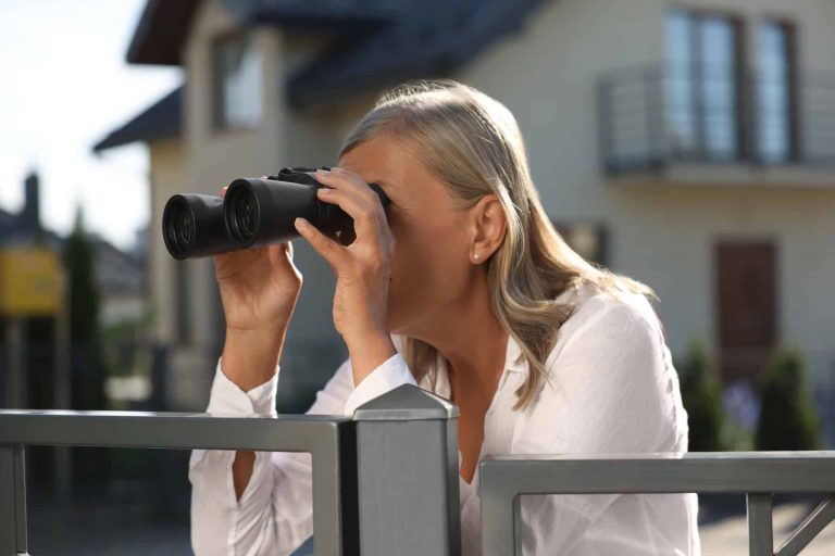Concept of private life. Curious senior woman with binoculars spying on neighbours over fence outdoors
