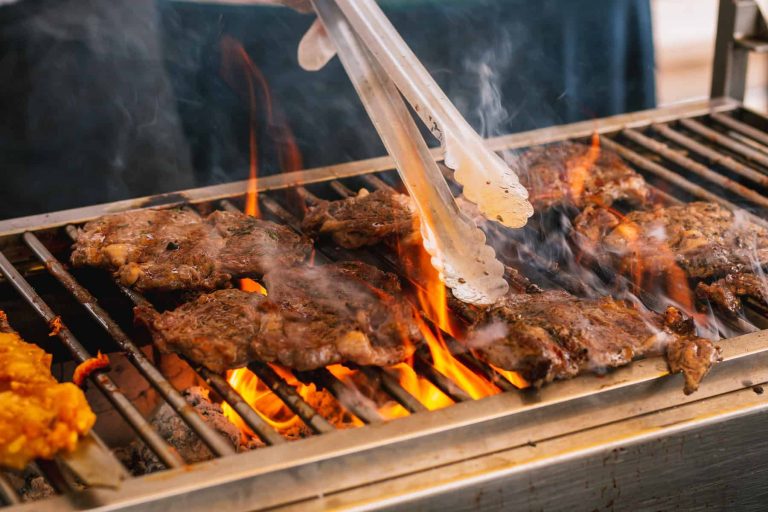 Serving tongs turning sizzling steaks on a grill with tongues of flame rising from the white charcoal