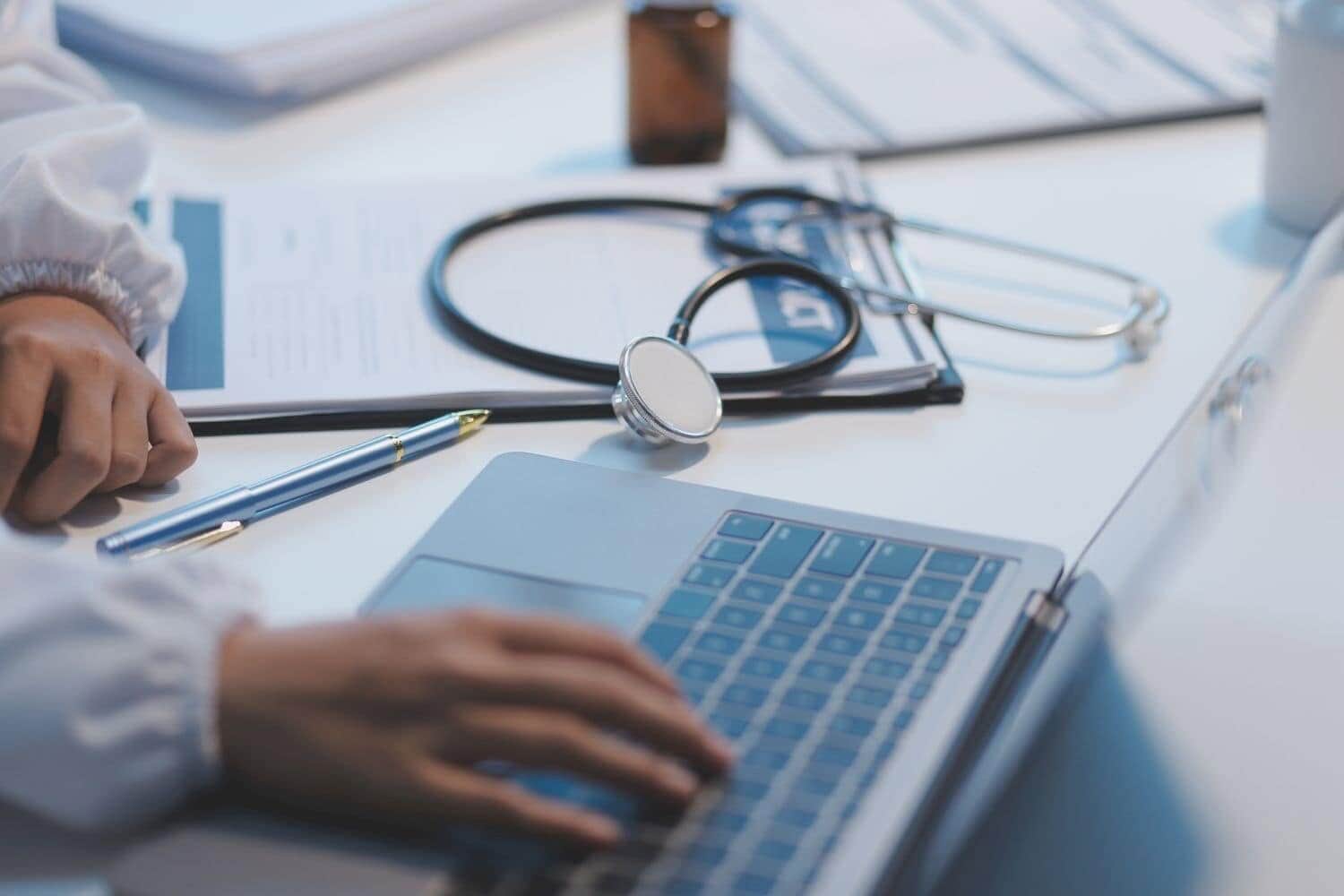 A professional and focused Asian female doctor in scrubs is working and reading medical research on her laptop in her office at a hospital.