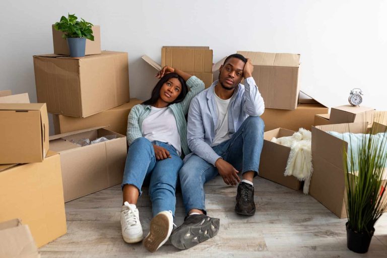 Sad black spouses exhausted after moving to new apartment, sitting among cardboard boxes and resting