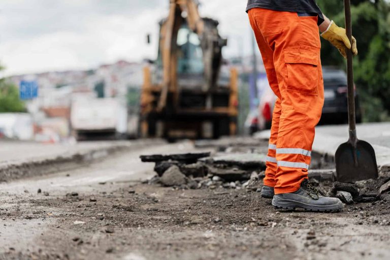 Construction worker repairs road surface with shovel while heavy machinery operates in background during cloudy day