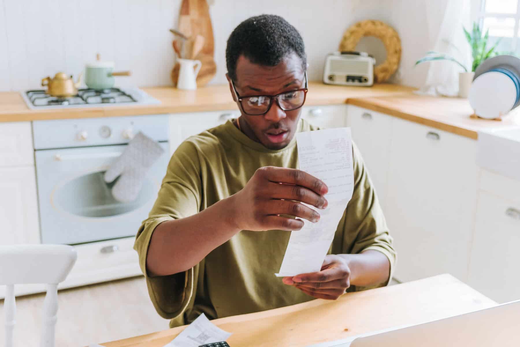 African American man in glasses, sitting at kitchen table, looks surprised as he reviews receipt, possibly indicating financial concern.