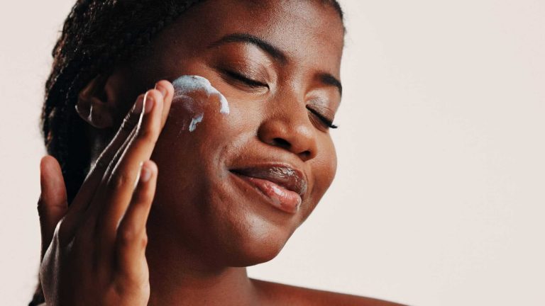 Happy, black woman and lotion on face in studio for facial hydration, treatment and sunscreen protection. Female person, apply cream and spf cosmetics for dermatology, anti aging and white background
