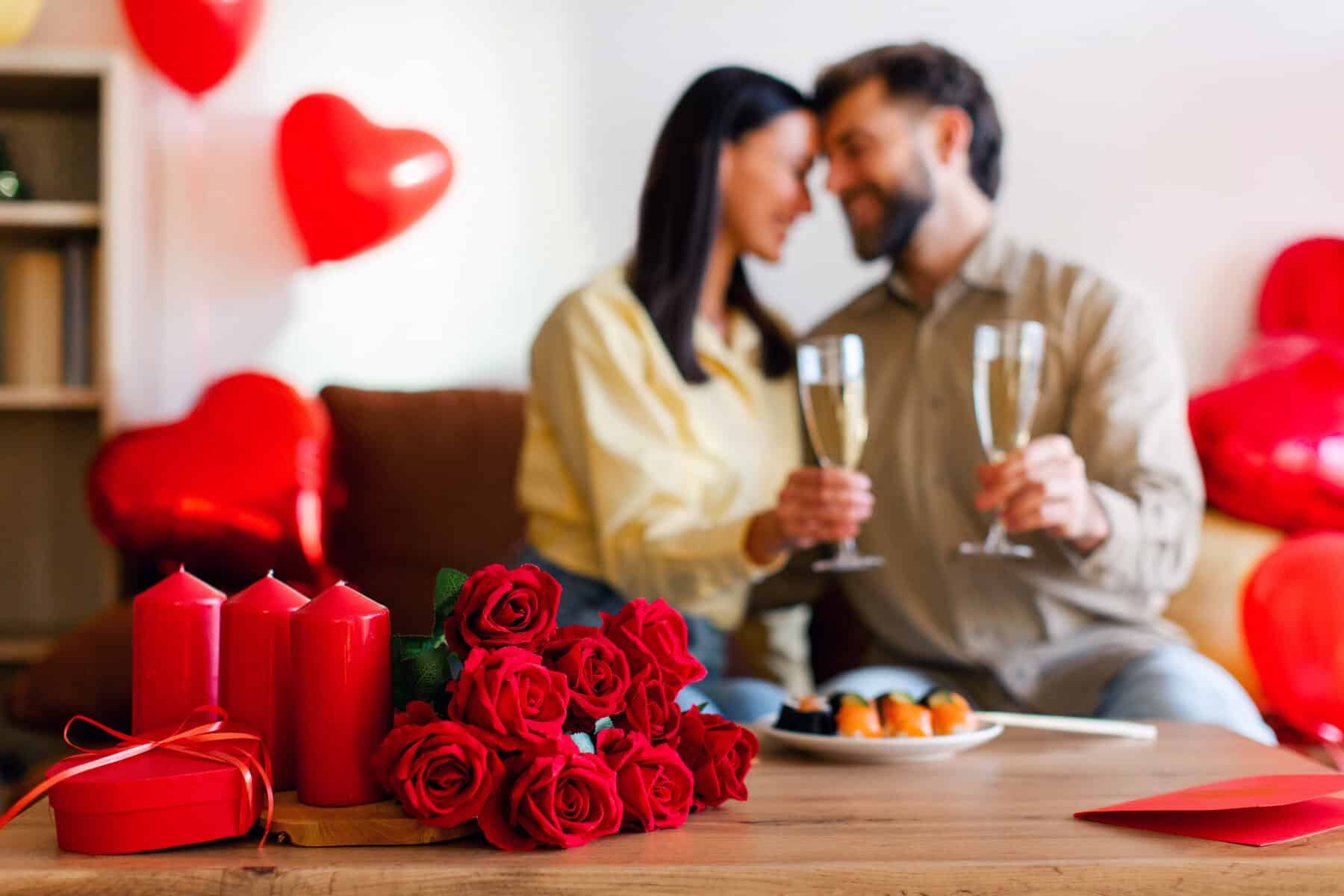 Focus on bouquet of red roses lying on table, happy man and woman spending time together at home, having romantic dinner in blurred background
