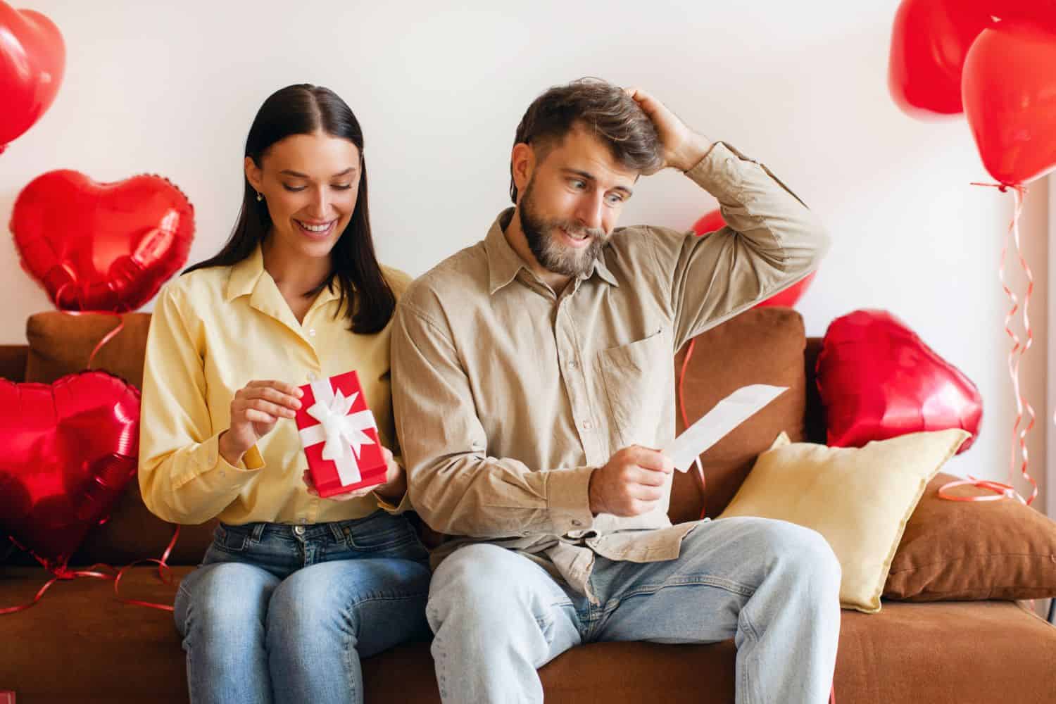 Happy woman looking at gift while man looking at bill, having problem with money, sitting on sofa in decorated room for Valentine's day