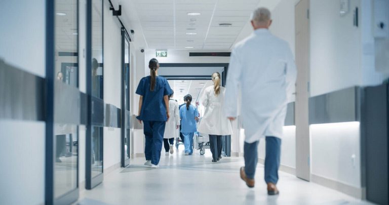 Hospital Hallway with Doctors, Nurses and Specialists in Hospital. Female and Male Physicians, Surgeons, Healthcare Officials Walk Together in Corridor with Their Back to Camera