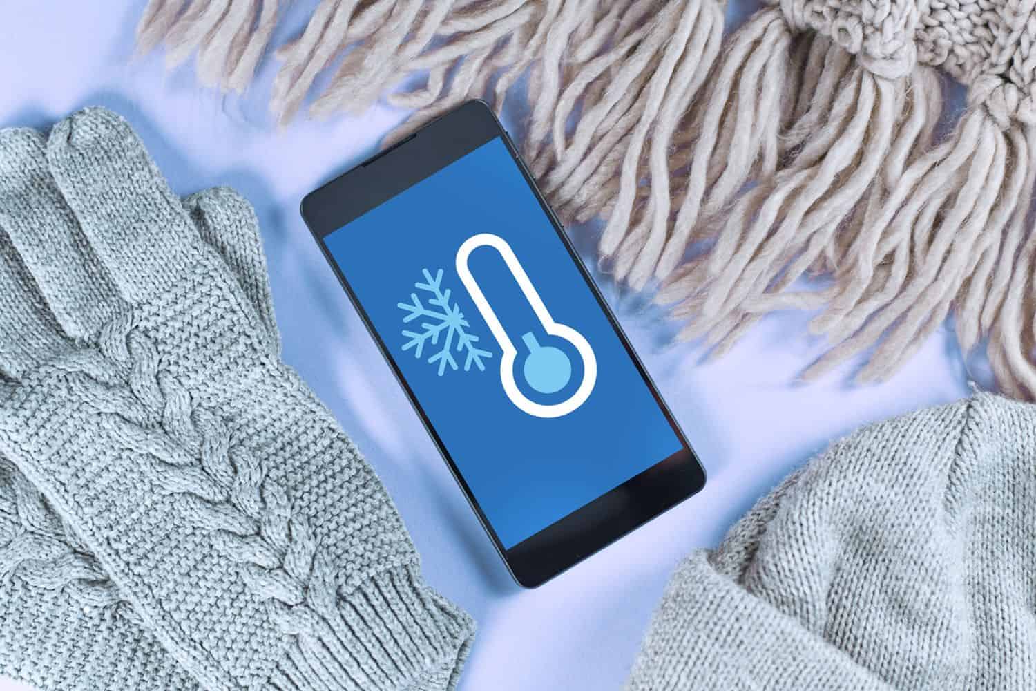 Concept for cold temperatures with snow and minus degrees with mobile phone showing weather forecast surrounded by warm clothes