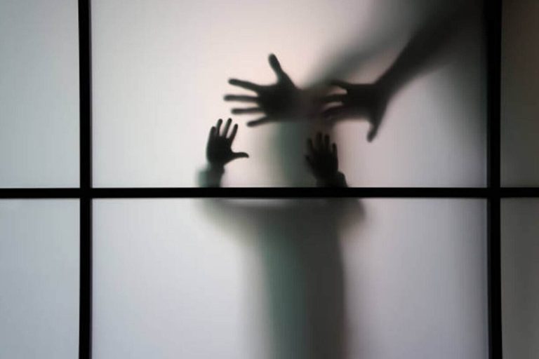 A woman stands behind of a frosted glass. She is afraid  of something.